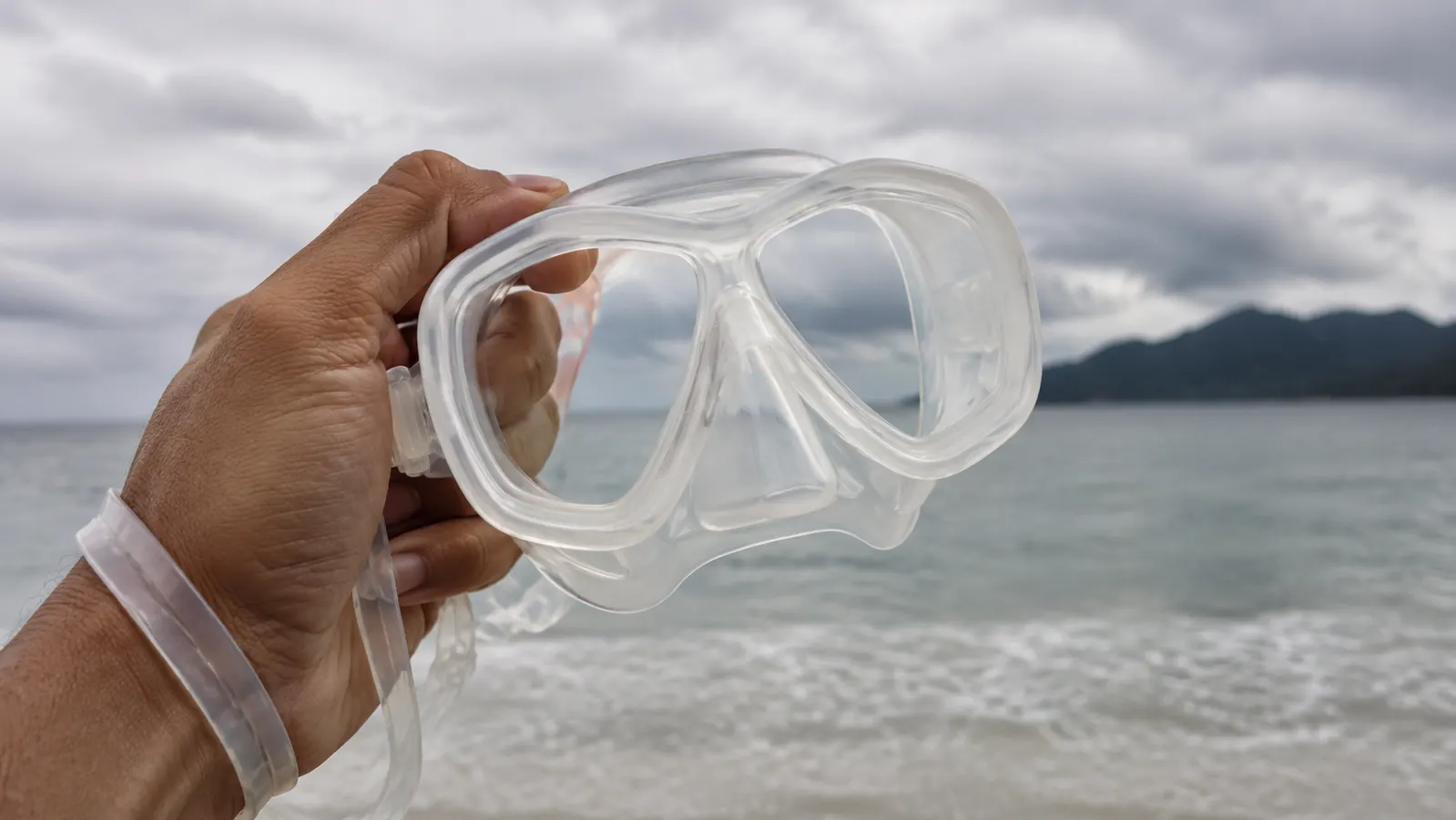 A weathered hand holding a frameless two-window dive mask by the skirt, strap tucked around the wrist, pre-dive on a beach with water behind