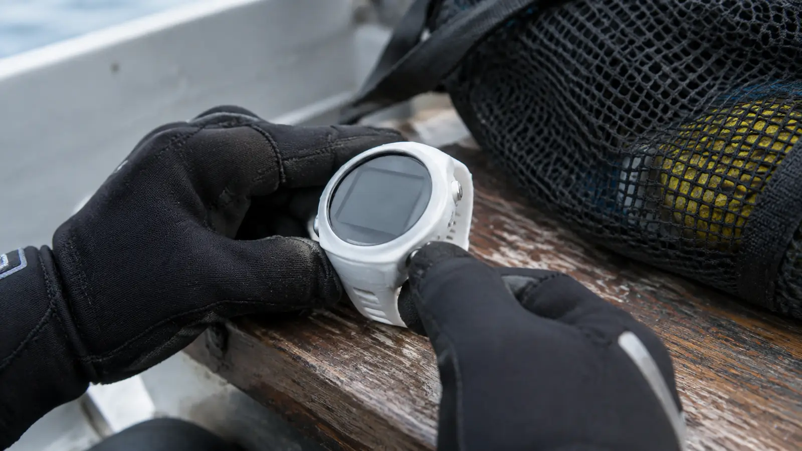 Hand in a neoprene glove pressing the dive computer's side button while it rests on a worn wooden boat bench