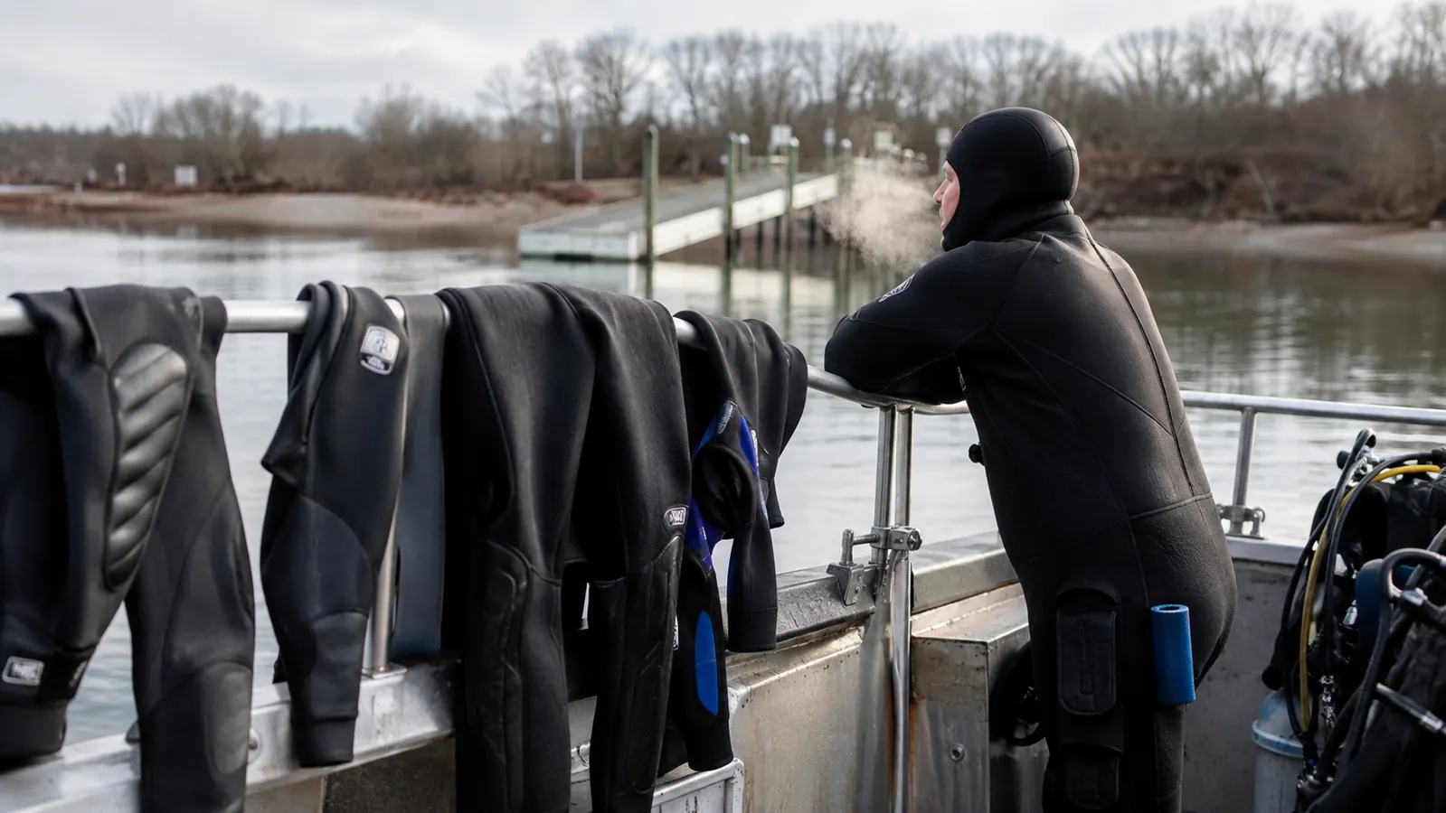 A short row of wetsuits hanging on the rail of a dive charter boat, a hooded diver leaning on the rail looking out at flat cold water, breath steaming, a bare treeline and launch ramp in the distance