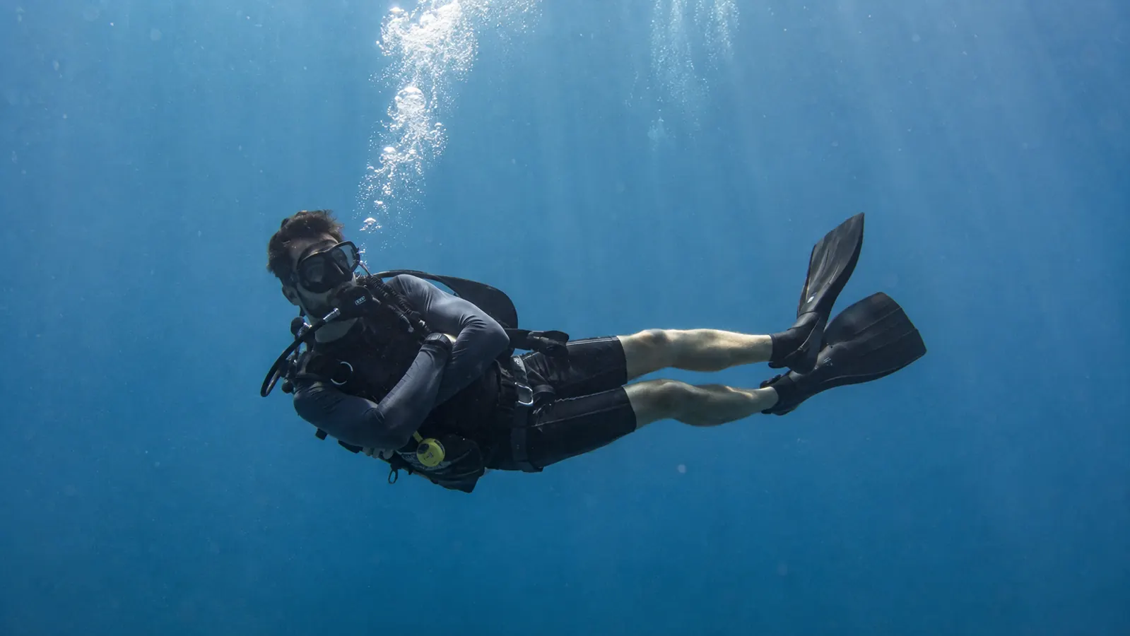 A scuba diver floats in relaxed horizontal trim at a safety stop, arms crossed loosely, bubbles rising from the regulator, deep blue water with faint sunbeams